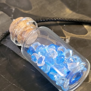 May include: A small, clear glass bottle pendant filled with blue and clear crystal beads. The bottle has a cork stopper and hangs from a black cord. The beads vary in shades of blue, creating a gradient effect.