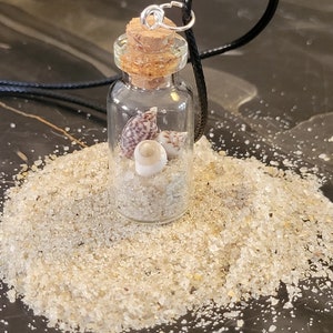 以下が含まれることがあります： コルク栓の小さなガラス瓶ネックレスで、砂と貝殻が入っています。瓶は白い砂の上に置かれています。