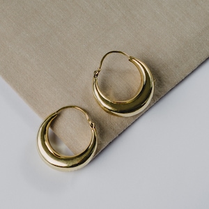 May include: A pair of gold hoop earrings with a curved, rounded shape. The earrings are on a beige fabric background.