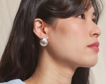 Può includere: Una donna indossa orecchini a forma di conchiglia d'argento. Gli orecchini sono di design semplice con una finitura liscia e lucida.