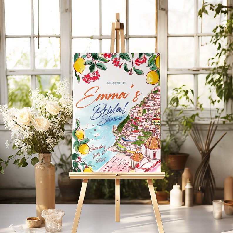 May include: A watercolor bridal shower sign on a wooden easel. The sign features the text "Welcome to Emma's Bridal Shower" with a coastal Italian town illustration, lemons, and the date "Saturday July 27, 2024".