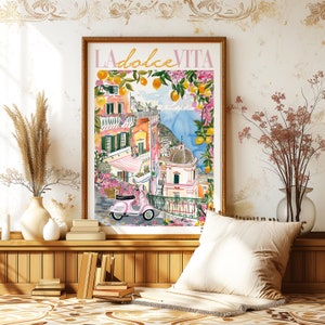 以下が含まれることがあります： 水彩画で描かれた、カラフルなイタリアの村の風景。手前にピンクのスクーターが描かれている。絵は額縁に入っていて、白い壁に飾られている。絵の上部にはピンクの文字で「LA dolce VITA」と書かれている。