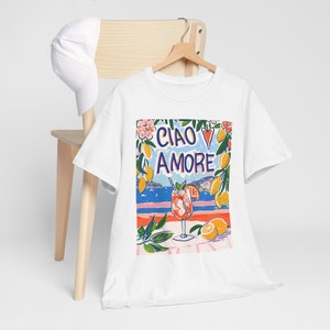May include: White t-shirt with a colorful graphic print of a glass of orange juice, lemons, flowers, and the text "Ciao Amore" with a red heart.