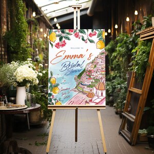 May include: A wooden easel displays a welcome sign for "Emma's Bridal Shower." The sign features a watercolor illustration of a coastal town with lemons and red flowers. The date "Saturday July 27, 2024" is also included.