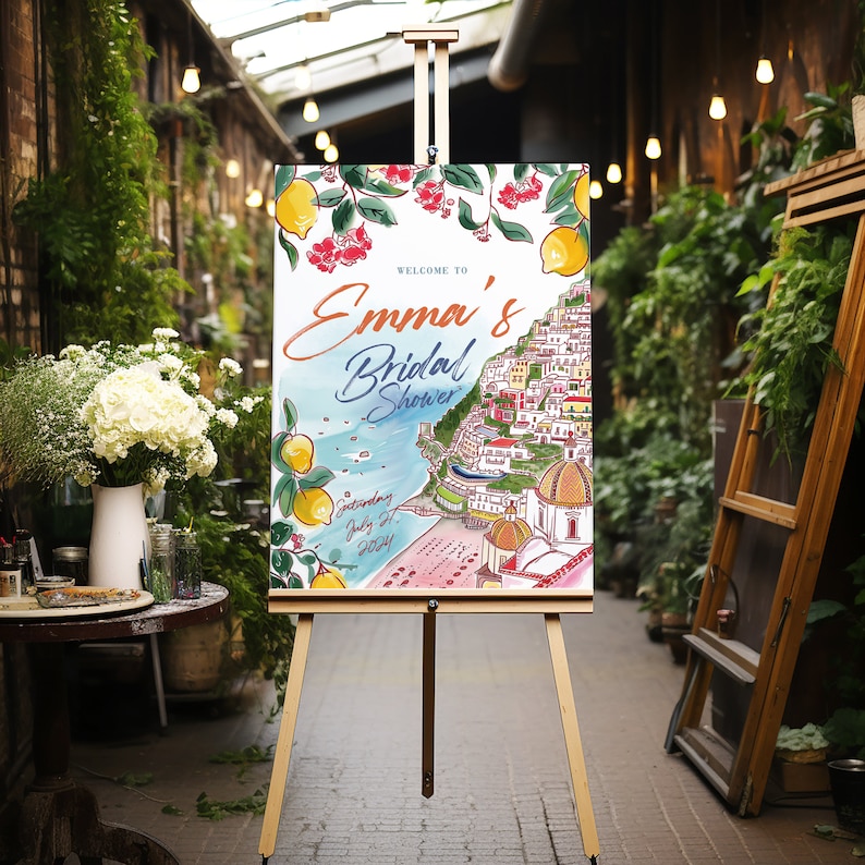 May include: A wooden easel displays a welcome sign for "Emma's Bridal Shower." The sign features a watercolor illustration of a coastal town with lemons and red flowers. The date "Saturday July 27, 2024" is also included.