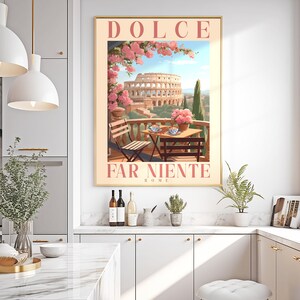 K&ouml;nnte beinhalten: Ein Vintage-Poster mit dem Text "Dolce Far Niente Rom" in rosa und wei&szlig;en Buchstaben. Das Poster zeigt eine farbenfrohe Illustration des Kolosseums in Rom mit einem Tisch und St&uuml;hlen auf einem Balkon mit Blick auf das Bauwerk. Rosa Blumen bl&uuml;hen an einer Rebe &uuml;ber dem Tisch.