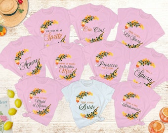 Puede incluir: Conjunto de diez camisetas rosas con varios diseños que incluyen cítricos naranjas, hojas verdes y texto. El texto en las camisetas incluye "That's Amore", "You Had Me At Spritz", "Cin Cin!", "Amore & Wishes for the Future Mrs!", "Prosecco Per Favore", "Here Comes La Sposa", "La Bella Sposa", "Come Si Dice Maid of Honor", "Come Si Dice Bride" y "Come Si Dice Bridesmaid".