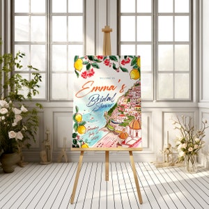 May include: A watercolor bridal shower sign on a wooden easel. The sign features the text "Welcome to Emma's Bridal Shower" with a coastal Italian town illustration, lemons, and red flowers. The date is Saturday, July 27, 2024.