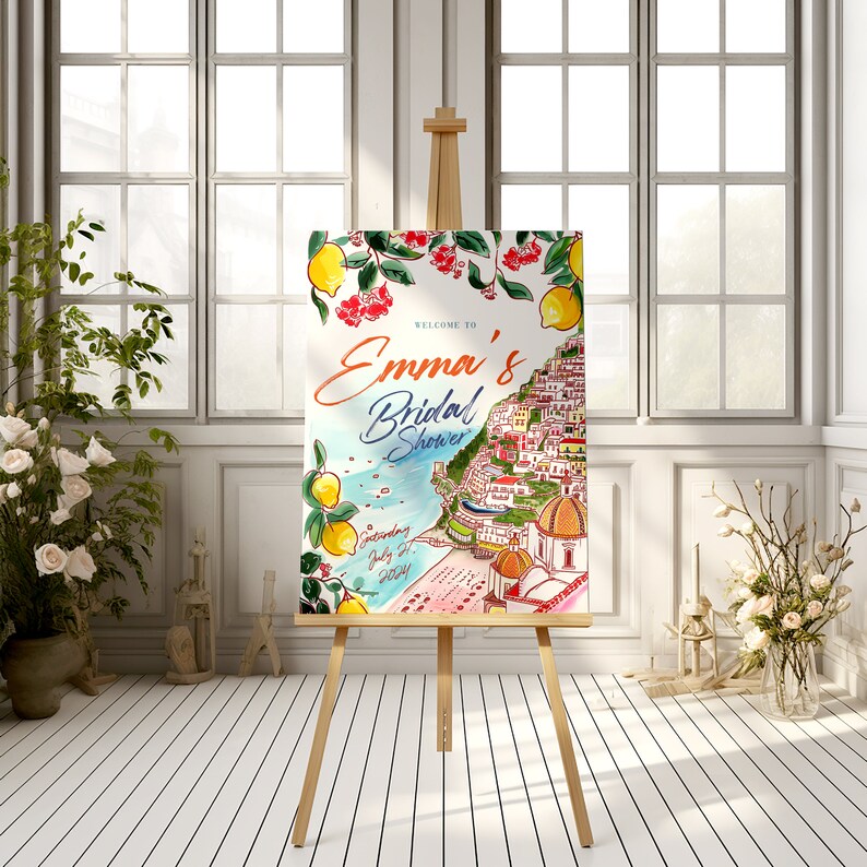 May include: A watercolor bridal shower sign on a wooden easel. The sign features the text "Welcome to Emma's Bridal Shower" with a coastal Italian town illustration, lemons, and red flowers. The date is Saturday, July 27, 2024.