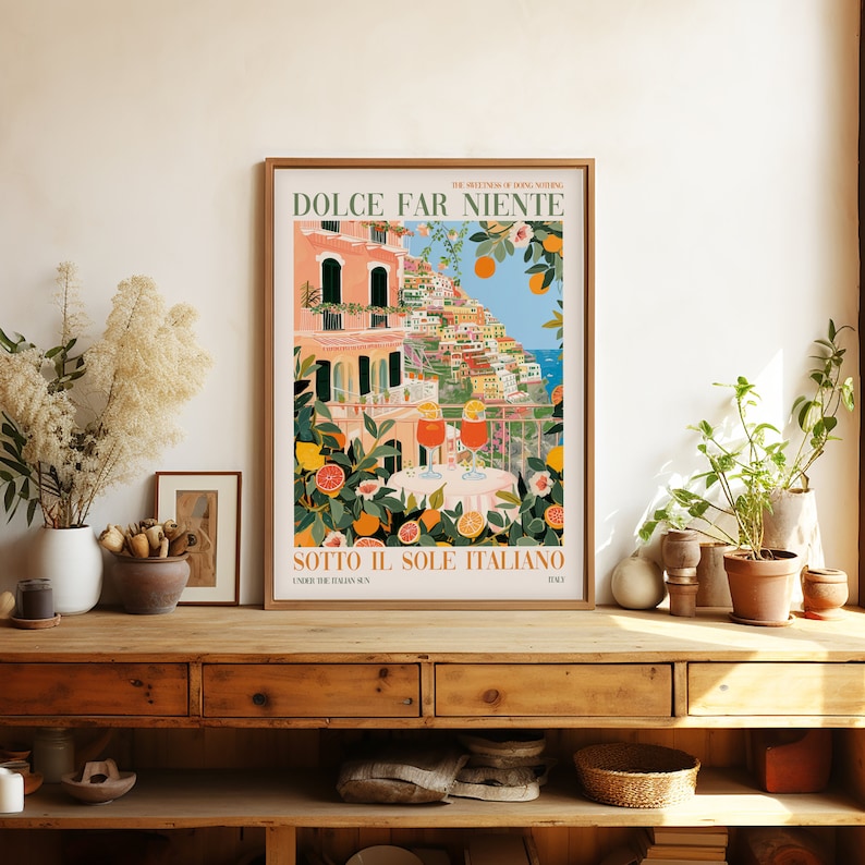 Op de afbeelding: Een kleurrijke poster met de tekst "Dolce Far Niente" en "Sotto Il Sole Italiano" in vintage stijl. De poster toont een sc&egrave;ne van een kleurrijk Italiaans dorp met een balkon met uitzicht op de oceaan. Twee glazen sinaasappelsap staan op een tafel met een bloemig tafelkleed.