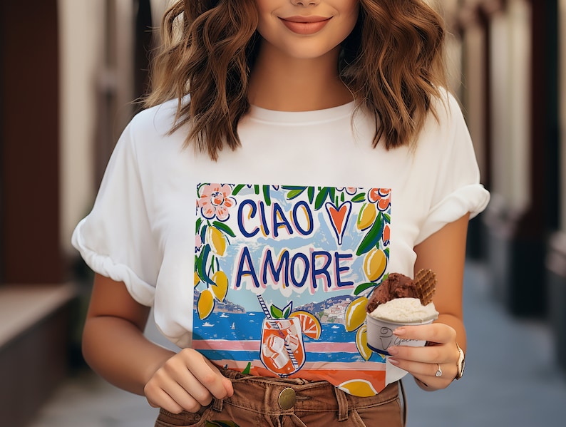 May include: White t-shirt with a colorful graphic print that says "Ciao Amore" with lemons, flowers, and a cocktail glass. The graphic also includes a blue and white striped pattern and a view of a city skyline.