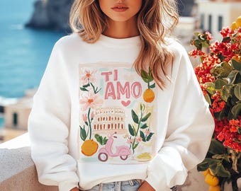 May include: White sweatshirt with a graphic print of a pink and white flower, a building, a scooter, and the text "Ti Amo" in pink letters.
