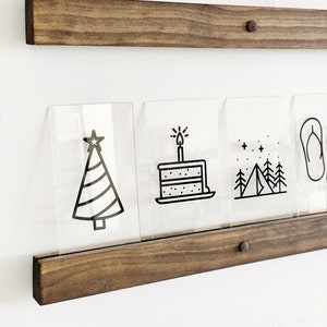 May include: A set of clear acrylic picture frames with black outlines of various icons, including a Christmas tree, a birthday cake, a camping scene, and a snowflake. The frames are displayed on a dark brown wooden shelf with a metal pin.