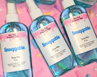 Könnte beinhalten: Mehrere transparente Body-Mist-Flaschen mit blauer Flüssigkeit sind auf einer rosa Oberfläche angeordnet. Jede Flasche hat ein weißes Etikett mit dem Text "Snuggable" und "Body Mist 4 oz." Die Etiketten enthalten auch den Markennamen "Nose Food Fragrance Shop."