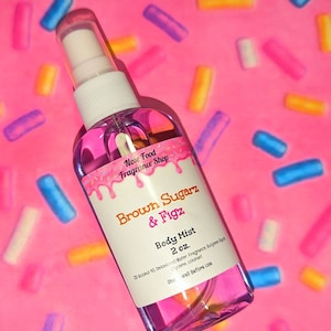 May include: A clear body mist spray bottle with a white label featuring the text "Brown Sugarz & Figz" and "Body Mist 2 oz." The bottle is filled with a pink liquid. The background is pink with colourful sprinkles.