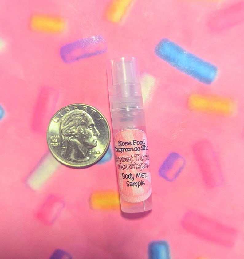 May include: A clear body mist sample bottle with a pink label that reads "Nose Food Fragrance Shop Sweet Tooth Boutique Body Mist Sample" next to a US quarter on a pink background with colorful sprinkles.