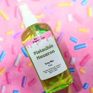 Puede incluir: Una botella de bruma corporal de vidrio transparente con un líquido verde y un pulverizador blanco. La etiqueta dice "Pistachio Macaron Body Mist 4 oz." con un diseño rosa y verde. El fondo es rosa con chispas de colores.