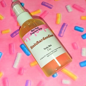 Puede incluir: Un frasco transparente de bruma corporal con una etiqueta blanca que dice "Snickerdoodles Body Mist 2 oz." La etiqueta tiene un diseño rosa y blanco. El frasco está sobre un fondo rosa con chispas de colores.