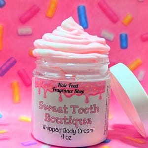 May include: A jar of pink whipped body cream from Sweet Tooth Boutique, topped with a swirl of pink cream. The label reads "Sweet Tooth Boutique Whipped Body Cream 4 oz." The background is pink with colorful sprinkles.