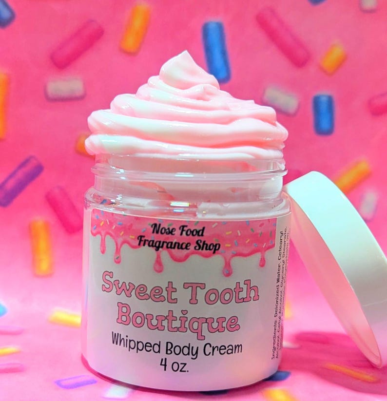 May include: A jar of pink whipped body cream from Sweet Tooth Boutique, topped with a swirl of pink cream. The label reads "Sweet Tooth Boutique Whipped Body Cream 4 oz." The background is pink with colorful sprinkles.