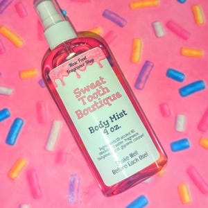 May include: A pink body mist spray bottle with a white label that reads "Sweet Tooth Boutique Body Mist 4 oz." The bottle is filled with a pink liquid and has a white spray nozzle. The background is pink with colorful sprinkles.