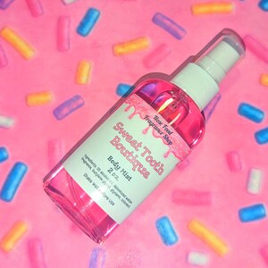 May include: A pink body mist spray bottle from Sweet Tooth Boutique, with a white label and cap. The label reads "Body Mist 2 oz." and "Nose Food Fragrance Shop." The background is pink with colorful sprinkles.