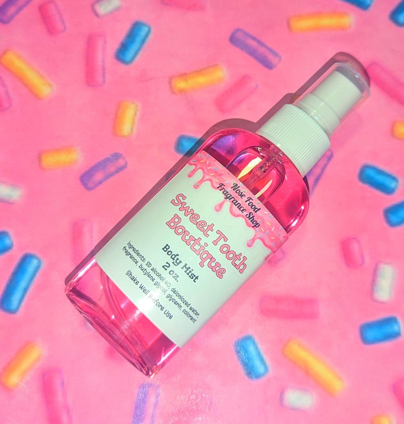 May include: A pink body mist spray bottle from Sweet Tooth Boutique, with a white label and cap. The label reads "Body Mist 2 oz." and "Nose Food Fragrance Shop." The background is pink with colorful sprinkles.