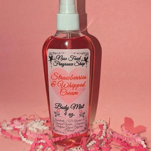 Puede incluir: Una botella de bruma corporal rosa con un pulverizador blanco. La etiqueta dice "Fresas y Crema Batida" y "Bruma Corporal 118 ml." La botella está rodeada de tiras de papel rosas y blancas y un recorte de mariposa rosa.