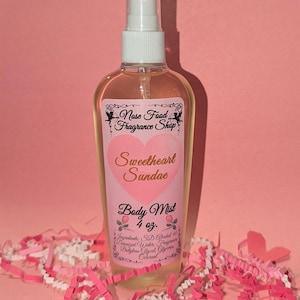 Puede incluir: Una botella transparente de bruma corporal con un pulverizador blanco y una etiqueta rosa. La etiqueta dice "Sweetheart Sundae" y "Body Mist 118 ml." La botella está rodeada de papel triturado rosa y blanco.