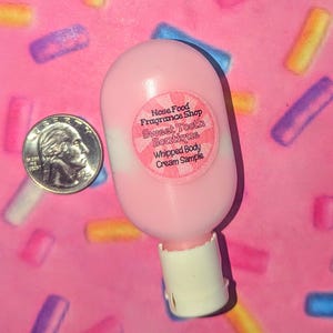May include: A pink whipped body cream sample bottle with a white base and a round sticker that reads "Nose Food Fragrance Shop Sweet Tooth Boutique Whipped Body Cream Sample." A U.S. quarter is next to the bottle.