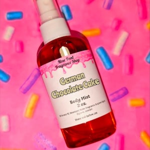 Puede incluir: Un frasco de bruma corporal con líquido rojo, etiquetado como "German Chocolate Cake". La etiqueta incluye el texto "Nose Food Fragrance Shop" y "2 oz". El frasco tiene una tapa blanca y está sobre un fondo rosa con chispas coloridas.