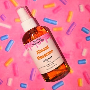 Puede incluir: Un frasco transparente de bruma corporal con una etiqueta blanca que dice "Almond Macaroon Body Mist 2 oz.". El frasco contiene un líquido dorado. El fondo es rosa con chispas de colores.