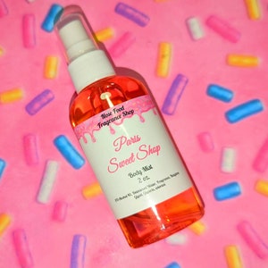 May include: A clear body mist spray bottle with a white label that reads "Paris Sweet Shop" and "Body Mist 2 oz." The liquid inside is orange. The bottle is set against a pink background with colorful sprinkles.