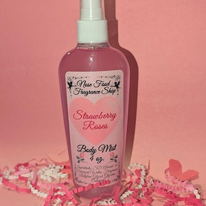 Puede incluir: Una botella de bruma corporal rosa con un pulverizador blanco. La etiqueta dice "Strawberry Roses Body Mist" y "4 oz." La botella está rodeada de papel triturado rosa y blanco y una mariposa de papel rosa sobre un fondo rosa.