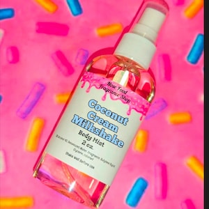 Puede incluir: Un frasco transparente de bruma corporal con un pulverizador blanco y una etiqueta que dice "Coconut Cream Milkshake Body Mist 2 oz." El frasco está sobre un fondo rosa con chispas de colores.