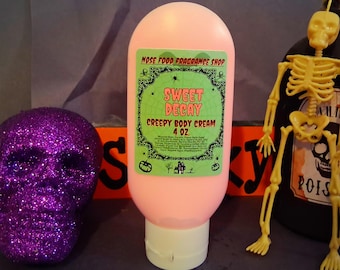 以下が含まれることがあります： 緑と黒のラベルが付いた、ピンク色の「Sweet Decay Creepy Body Cream」のボトル。 ラベルには「Nose Food Fragrance Shop」と「4 oz」の文字が含まれています。 紫色のグリッタースカルと黄色のスケルトンが背景にあります。