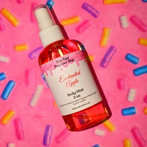 Puede incluir: Botella transparente de bruma corporal con un pulverizador blanco, llena de líquido rojo. La etiqueta dice "Enchanted Apple Body Mist 2 oz.". Fondo rosa con confeti colorido.