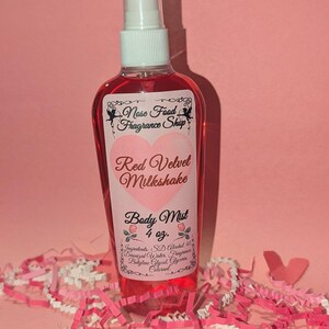 Puede incluir: Una botella de bruma corporal roja con un pulverizador blanco. La etiqueta dice "Red Velvet Milkshake Body Mist 4 oz." con el logo "Nose Food Fragrance Shop". La botella está rodeada de papel triturado rosa y blanco sobre un fondo rosa.