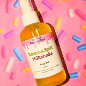 Puede incluir: Un frasco de spray transparente lleno de líquido amarillo, etiquetado como "Banana Split Milkshake Body Mist" de Nose Food Fragrance Shop. El fondo es rosa con chispas de colores.