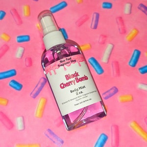 Puede incluir: Un frasco de spray transparente lleno de líquido rosa, etiquetado como "Black Cherry Bomb Body Mist, 2 oz." El frasco tiene una tapa blanca y una etiqueta con detalles rosados goteando. El fondo es rosa con chispas coloridas.