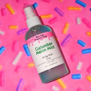 Puede incluir: Un frasco transparente de bruma corporal con tapa blanca y etiqueta que dice "Cucumber Melon Mint". La etiqueta tiene detalles rosas goteando y el texto "Nose Food Fragrance Shop". El frasco es de 59 ml y el fondo es rosa con confeti colorido.