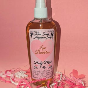Puede incluir: Una botella transparente de bruma corporal con una etiqueta rosa con un diseño de corazón y las palabras "Luv Bubbles". La botella contiene un líquido dorado y está etiquetada como "Body Mist 4 oz". El fondo es rosa con confeti de papel rosa y blanco.