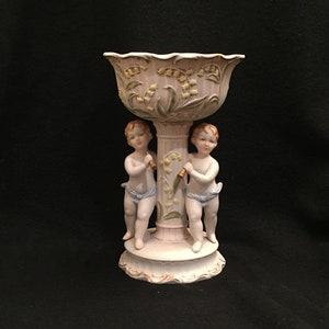 Puede incluir: Un tazón de porcelana blanca con un diseño floral y dos figuras de querubines en la base. El tazón está decorado con hojas verdes y flores amarillas.