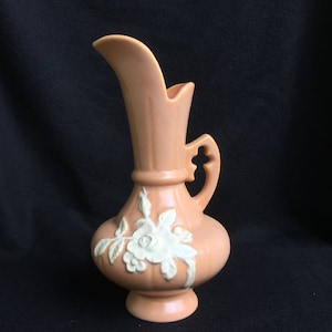 Puede incluir: Una jarra de cerámica de color melocotón con un diseño floral blanco. La jarra tiene una boca ancha y un asa en el lateral.