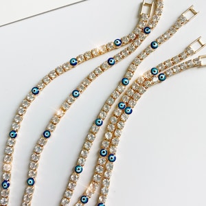 May include: Close-up of several gold-toned tennis bracelets with clear, sparkling stones. Each bracelet features small, blue evil eye charms. The bracelets are arranged on a white surface, with the Instagram handle @FRESHALAMODE visible.