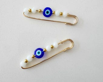 Puede incluir: Dos imperdibles de oro con cuentas blancas y un amuleto de ojo turco azul. Los amuletos son redondos con un centro blanco y un anillo exterior azul.