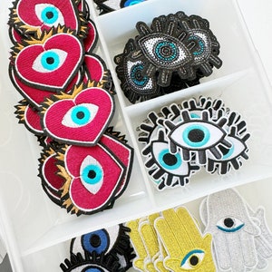 May include: Assortment of colorful fabric patches featuring the evil eye symbol in various shapes, including hearts, hands, and traditional eye shapes. The patches are arranged in a clear plastic storage container.