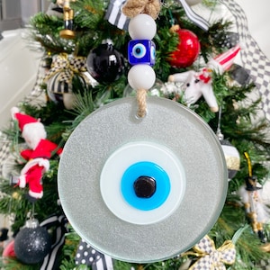 May include: A round, silver-colored ornament with a blue evil eye design. The ornament is hanging from a brown twine rope with white and blue beads.