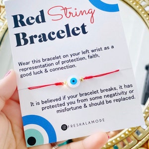 May include: Red string bracelet with a blue and white evil eye charm. The bracelet is tied around a wrist and the card reads "Red String Bracelet. Wear this bracelet on your left wrist as a representation of protection, faith, good luck & connection. It is believed if your bracelet breaks, it has protected you from some negativity or misfortune & should be replaced. FRESHALAMODE"