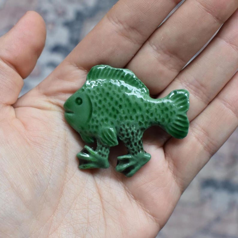Puede incluir: Una peque&ntilde;a figura de pez de cer&aacute;mica en color verde bosque, con acabado brillante y una superficie texturizada que imita escamas. El pez presenta una aleta dorsal prominente, una cola definida y dos pies palmeados distintos.